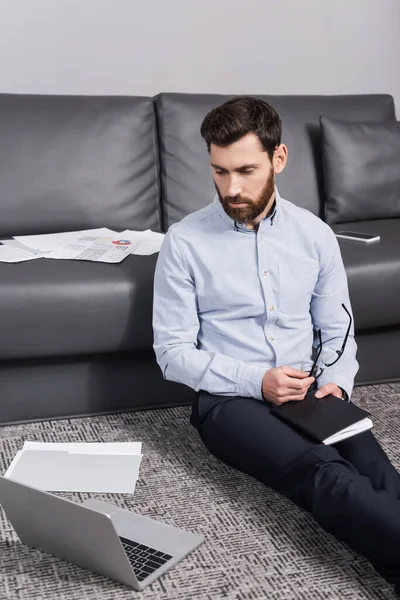 Bearded freelancer holding eyeglasses and notebook near laptop and couch with documents — Stock Photo