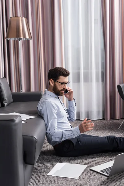 Barbuto freelance in occhiali seduto su tappeto e parlando su smartphone vicino laptop e divano — Foto stock