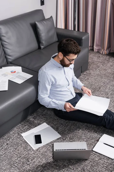 Visão de alto ângulo de freelancer barbudo em óculos sentado no tapete perto de documentos e gadgets — Fotografia de Stock