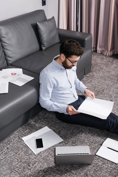 Vista de ángulo alto del freelancer barbudo en gafas sentadas en la alfombra cerca de documentos y gadgets - foto de stock