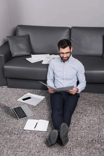 Cheerful freelancer in eyeglasses sitting on carpet and looking at folder near gadgets — Stock Photo