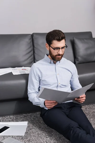 Bearded man in eyeglasses sitting on carpet and looking at folder near gadgets — Stock Photo