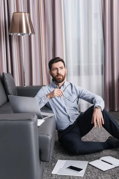 Hombre pensativo y barbudo sosteniendo anteojos y pensando cerca de gadgets en habitación de hotel - foto de stock