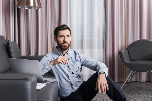 Pensive and bearded man holding eyeglasses and thinking near laptop on sofa — Stock Photo