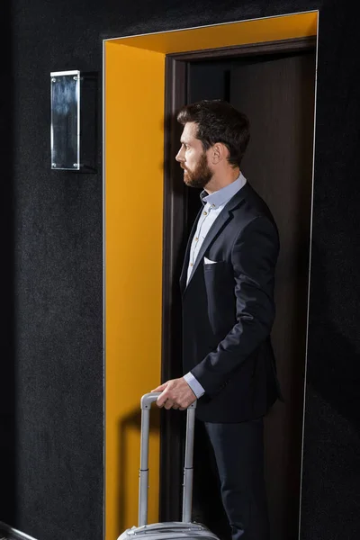 Hombre barbudo de traje de pie con equipaje en el pasillo del hotel - foto de stock