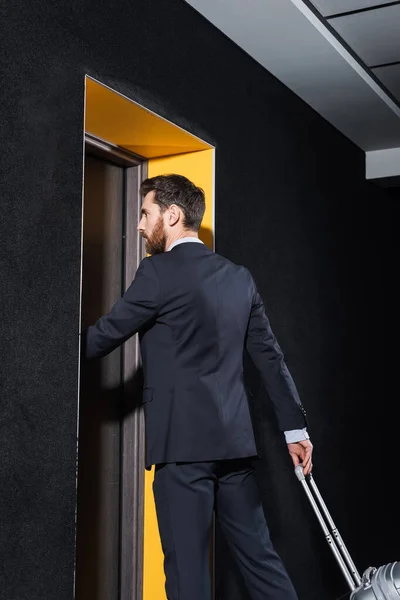 Vista lateral del hombre barbudo en traje con asa de equipaje al entrar en la habitación del hotel - foto de stock