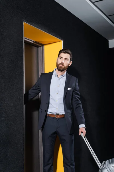 Uomo barbuto in giacca e cravatta in possesso di maniglia bagagli guardando lontano nel corridoio dell'hotel — Foto stock