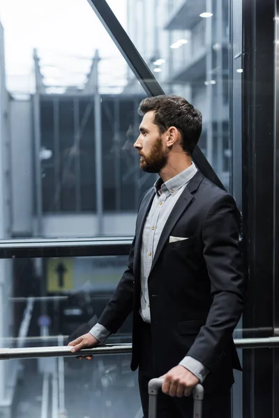 Vue latérale de l'homme barbu en costume tenant poignée de bagages dans l'ascenseur moderne — Photo de stock