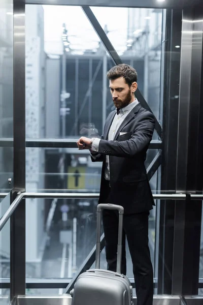 Uomo barbuto in tuta tempo di controllo sul orologio da polso vicino ai bagagli in ascensore moderno — Foto stock