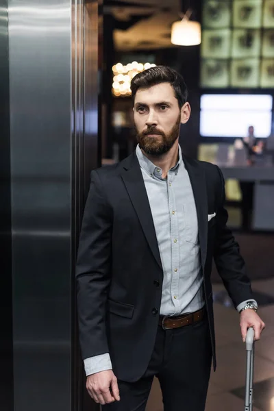 Hombre de negocios barbudo en traje con asa de equipaje en ascensor moderno - foto de stock