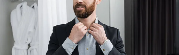 Vista recortada del hombre alegre en traje ajustando el cuello de la camisa en la habitación de hotel, bandera — Stock Photo