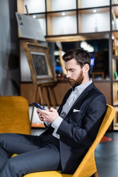 Hombre barbudo de traje usando teléfono inteligente en el vestíbulo del hotel - foto de stock