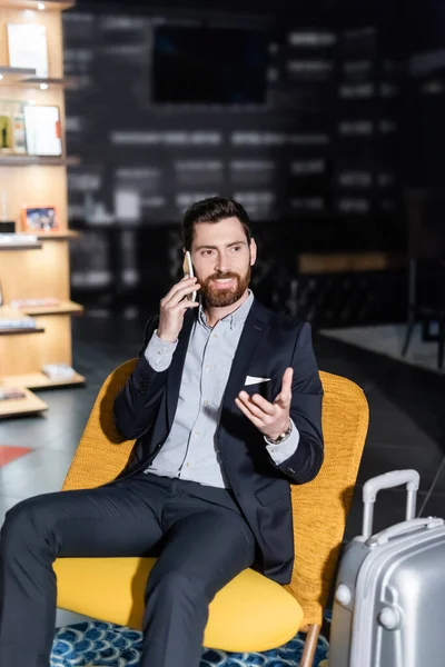 Hombre feliz en traje hablando en el teléfono inteligente cerca del equipaje en el vestíbulo del hotel - foto de stock