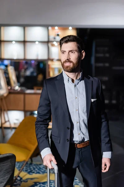 Homme barbu en costume tenant poignée de bagages dans le foyer de l'hôtel — Photo de stock