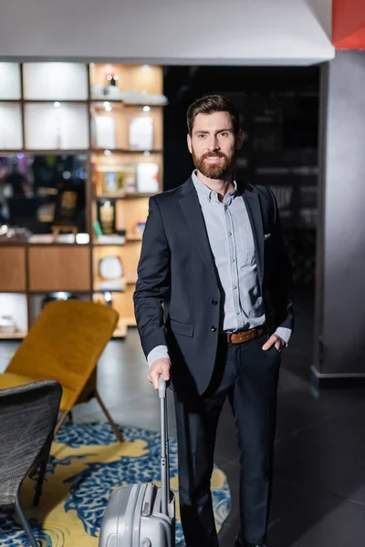 Cheerful man in suit holding luggage handle and posing with hand in pocket in hotel foyer — Stock Photo