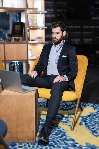 Homme barbu avec badge d'identité regardant ordinateur portable tout en étant assis dans le foyer de l'hôtel — Photo de stock