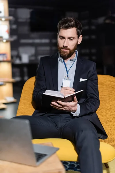 Bärtiger Mann mit Ausweis schreibt auf Notizbuch, während er im Hotelfoyer einen verschwommenen Laptop benutzt — Stockfoto