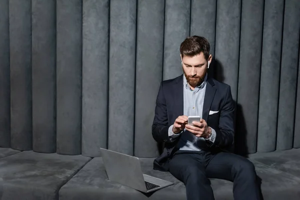 Bearded man in earphones using smartphone near laptop in hotel lobby — Stock Photo