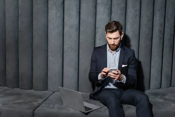 Uomo barbuto in auricolare utilizzando smartphone vicino al computer portatile nella hall dell'hotel — Foto stock