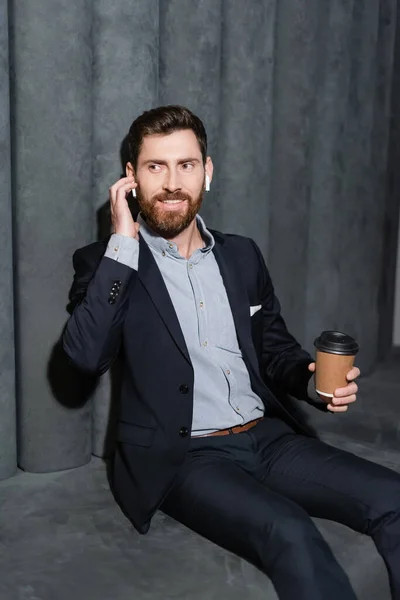 Happy businessman in earphones and suit holding paper cup in hotel foyer — Stock Photo