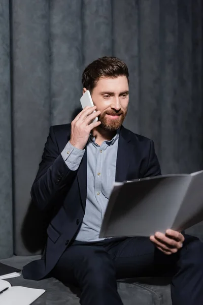 Hombre alegre y barbudo mirando la carpeta mientras habla en el teléfono celular en el vestíbulo del hotel - foto de stock
