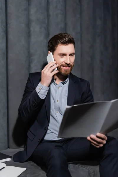 Fröhlicher und bärtiger Mann schaut in Hotellobby auf Ordner, während er mit dem Handy telefoniert — Stockfoto