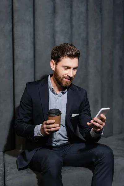 Hombre sonriente en traje usando teléfono inteligente y sosteniendo la taza de papel en el vestíbulo del hotel - foto de stock
