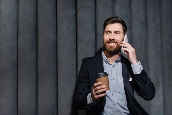 Hombre alegre en traje hablando en el teléfono inteligente y sosteniendo taza de papel en el vestíbulo del hotel - foto de stock
