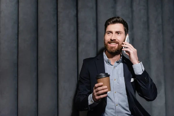 Cheerful man in suit talking on smartphone and holding paper cup in hotel lobby — Stock Photo