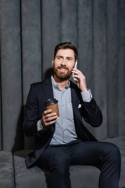 Alegre hombre de negocios en traje hablando en el teléfono inteligente y sosteniendo taza de papel en el vestíbulo del hotel - foto de stock