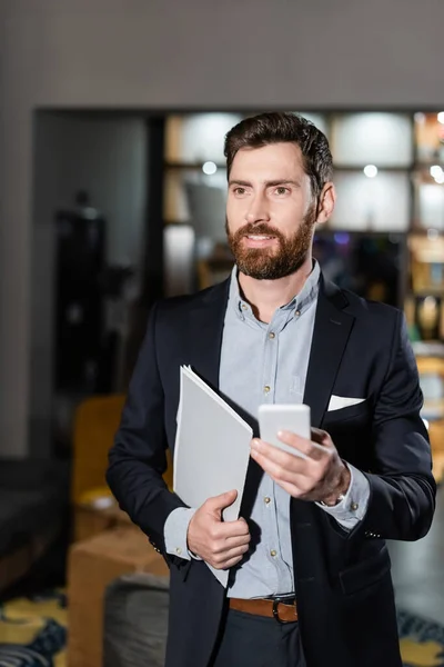 Cheerful man in suit holding folder and mobile phone in hotel lobby — Stock Photo