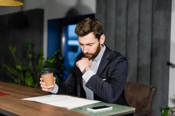 Thoughtful man in suit holding paper cup while looking at documents in hotel cafe — Stock Photo