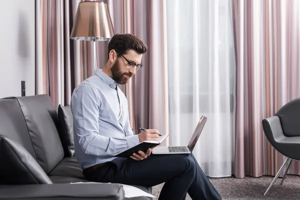 Homme en chemise et lunettes écriture sur ordinateur portable près de portable dans la chambre d'hôtel — Photo de stock