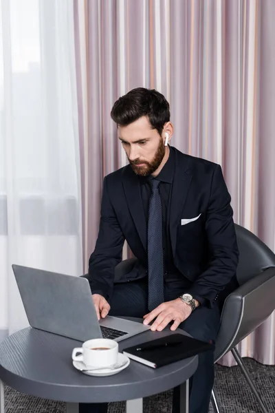 Uomo barbuto in auricolare senza fili utilizzando il computer portatile in camera d'albergo moderno — Foto stock