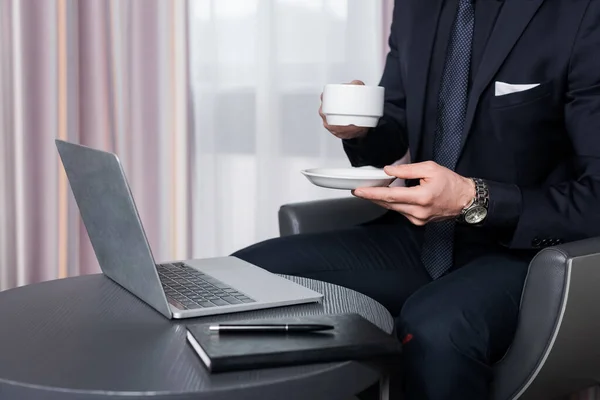 Vista recortada del hombre en traje que sostiene la taza y el platillo cerca del ordenador portátil en la habitación de hotel moderna - foto de stock