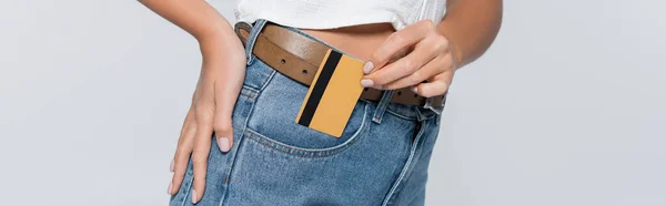 Vista ritagliata di giovane donna che mette carta di credito con cashback in tasca su jeans isolati su grigio, banner — Foto stock