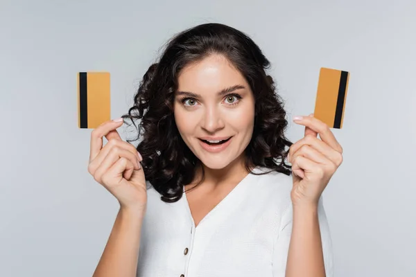 Здивована молода жінка тримає кредитні картки з готівкою ізольовані на сірому — стокове фото