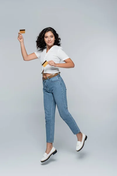 Full length of cheerful young woman in jeans holding credit cards with cashback while levitating on grey — Stock Photo