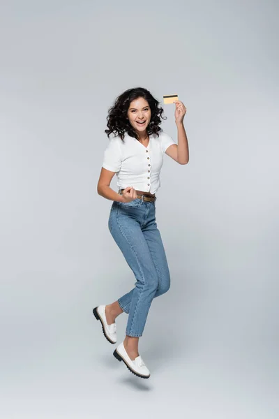 Полная длина веселой молодой женщины в джинсах, держащей кредитную карту с возвратом денег, в то время как левитация на серой — стоковое фото
