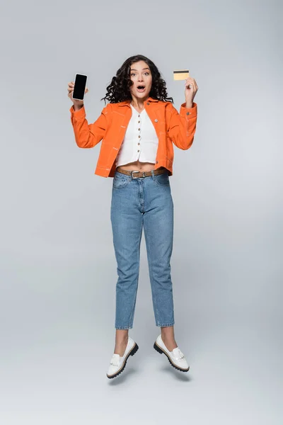 Full length of shocked woman in orange jacket holding credit card with cashback and smartphone while levitating on grey — Stock Photo