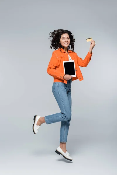 Full length of happy woman in orange jacket holding credit card with cashback and digital tablet while levitating on grey — Stock Photo