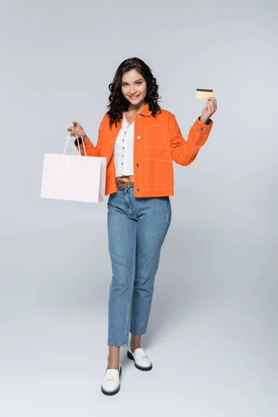 Полная длина счастливой женщины в оранжевой куртке, держащей кредитную карту с возвратом денег и сумку с покупками на сером — стоковое фото