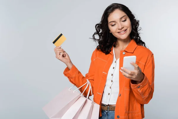 Mujer feliz usando teléfono inteligente mientras sostiene la tarjeta de crédito con cashback y bolsas de compras aisladas en gris - foto de stock