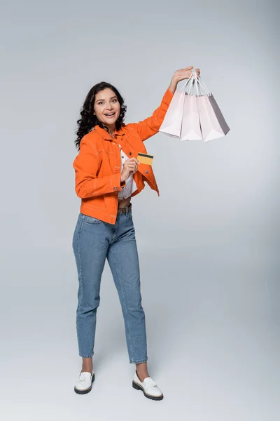 Повна довжина радісної молодої жінки, що тримає кредитну картку з готівкою та сумками на сірому — стокове фото