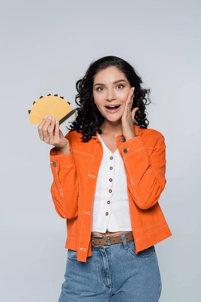 Mujer joven sorprendida en chaqueta naranja con tarjetas de crédito con cashback aislado en gris - foto de stock