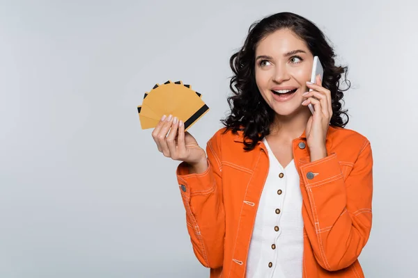 Mujer joven feliz en chaqueta naranja con tarjetas de crédito con cashback y hablando en el teléfono inteligente aislado en gris - foto de stock
