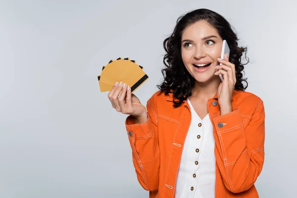 Happy young woman in orange jacket holding credit cards with cashback and talking on smartphone isolated on grey — Stock Photo