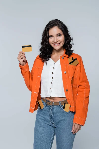 Счастливая молодая женщина с кредитными картами в карманах джинсов и оранжевой куртки изолированы на серый — стоковое фото