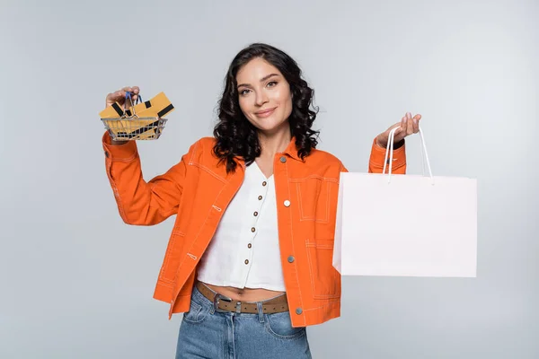 Junge Frau in orangefarbener Jacke hält Einkaufskorb mit Kreditkarten und Papiertüte isoliert auf grau — Stock Photo
