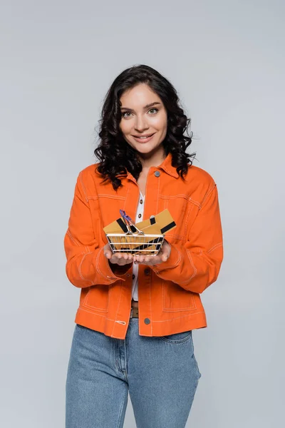 Mujer joven positiva en chaqueta naranja sosteniendo cesta de la compra con tarjetas de crédito aisladas en gris - foto de stock